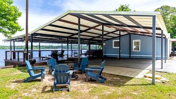 A covered outdoor seating area with Adirondack chairs, a grill, and a view of a lake.