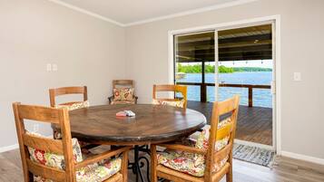 Un coin repas avec une table ronde en bois et des chaises, une vue sur un lac par une porte vitrée, et une terrasse en bois à l’extérieur.