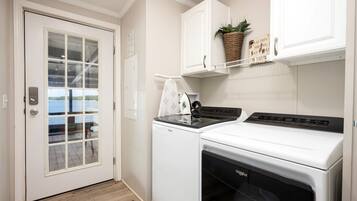 A laundry room with two washing machines, a drying machine, a window with a view of a body of water, and a cabinet with a plant and a sign.
