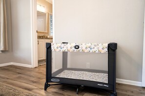 Een Graco Pack 'n Play kinderbedje met een patroon bovenop, staat op een houten vloer in een kamer met een witte deur en een keuken op de achtergrond.