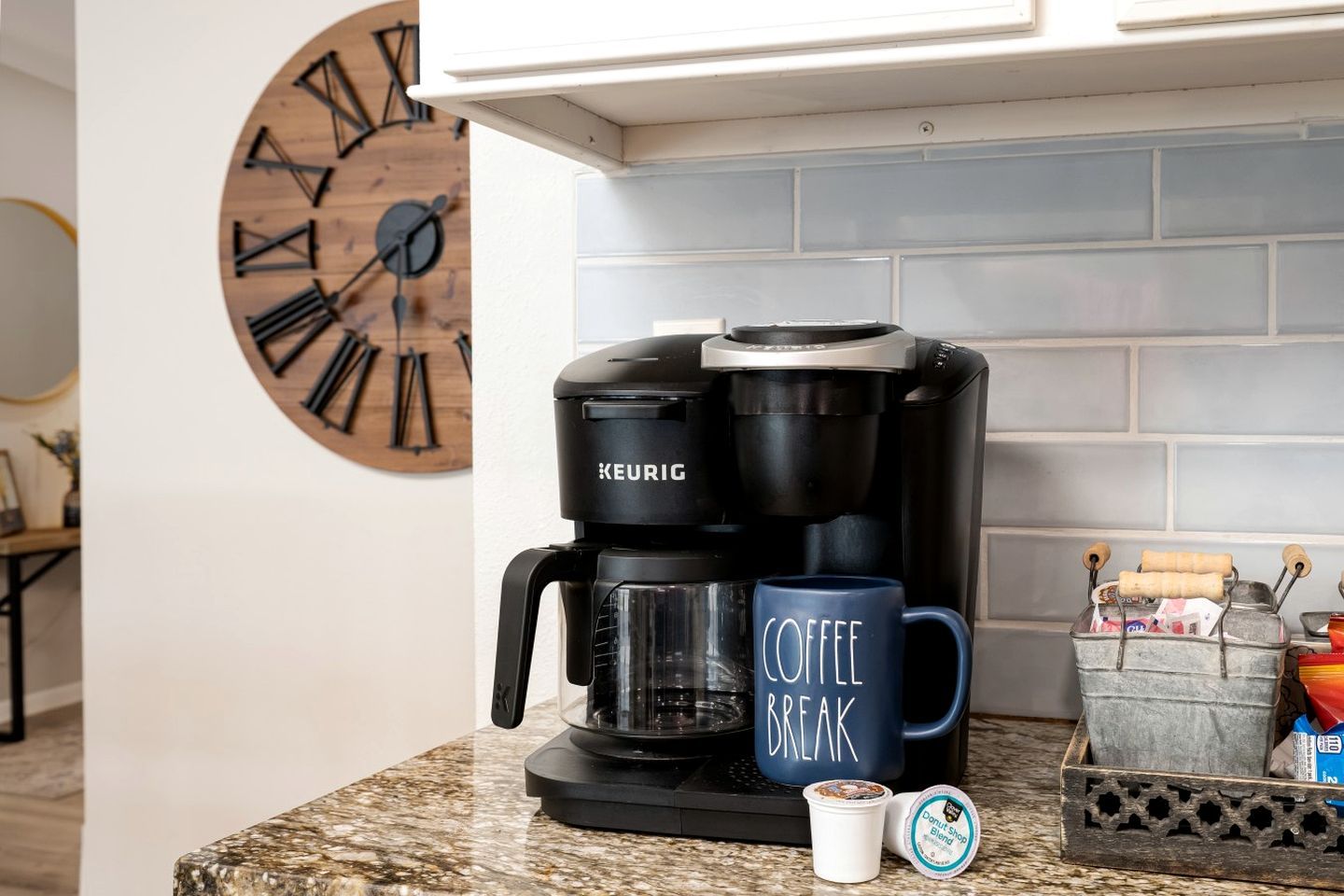 Una cafetera Keurig, una taza con la leyenda 'Coffee Break', una taza pequeña de café y un recipiente con cápsulas de café sobre una encimera de cocina.