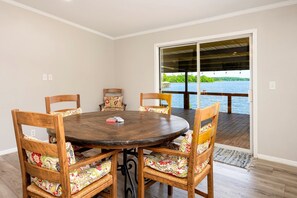 Un coin repas avec une table ronde en bois et des chaises, une vue sur un lac par une porte vitrée, et une terrasse en bois à l’extérieur.