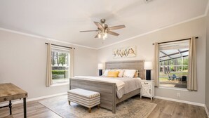 Un dormitorio con cama, mesita de noche, banco, ventilador de techo y ventanas grandes.