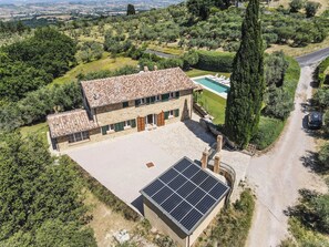Une maison en pierre avec une piscine, entourée de verdure et bordée d’une route.
