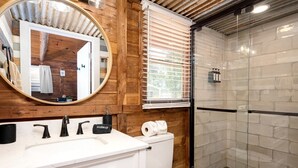 A bathroom with a wooden wall, a round mirror, a white sink, a toilet, and a glass shower enclosure. - Rustic Waterfront Cabin on Toledo Bend Lake for an Unforgettable Getaway in Nature in Hemphill, Texas (Hemphill)