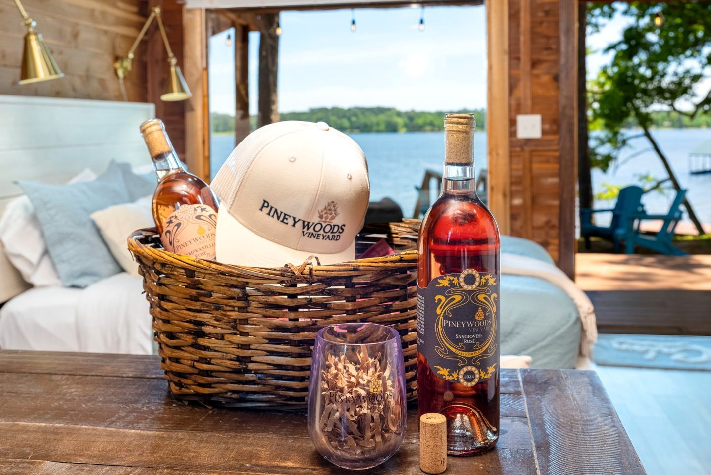 A wicker basket with a Piney Woods Vineyard cap, a bottle of Sangivose Rose wine, and a wine glass on a wooden table.