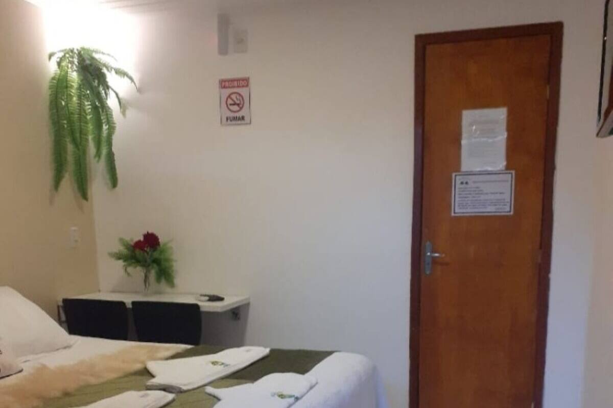Quarto de hotel com uma cama, uma pequena mesa com uma planta, uma porta com placa e piso de azulejos.