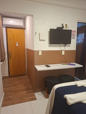 Habitación de hotel con puerta de madera, televisor montado en la pared, estante blanco con objetos, cama con manta blanca y taburete negro.