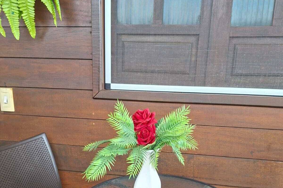 Mesa de vidro redonda com um vaso branco contendo uma rosa vermelha e folhagem verde, colocada entre duas cadeiras de vime, em frente a uma parede de madeira com uma janela.