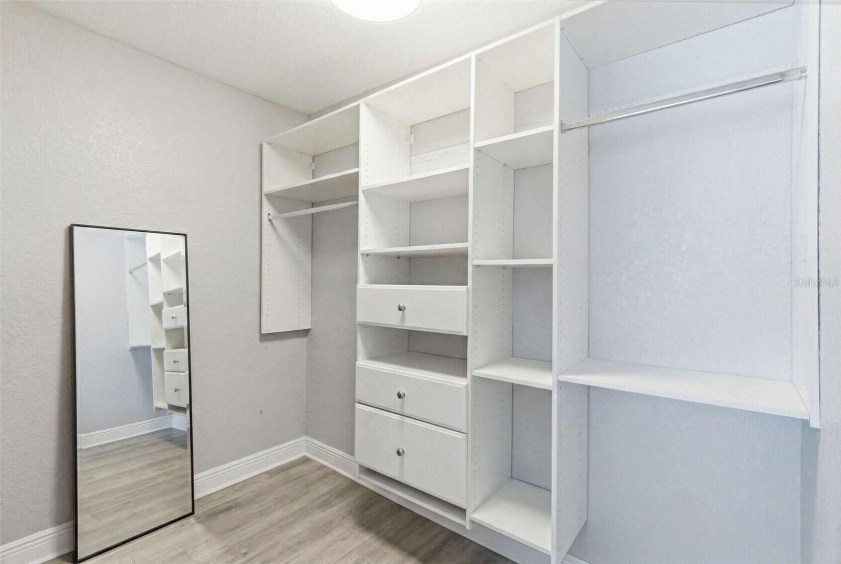 Um closet espaçoso com prateleiras abertas, porta espelhada e piso de madeira.
