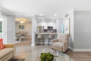 A modern living room with a glass coffee table, a sofa, a wicker armchair, and a dining area with a table and chairs. - Spacious 3BR Condo in Gated St. Petersburg Community with Private Beach (St. Petersburg)