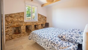 A bedroom with a stone fireplace, a bed with floral bedding, and a wooden ceiling beam. - Between Vineyards & the Sea | Beaches Within Walking Distance | Air Conditioning & Wi-Fi (La Ciotat)