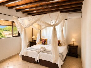 Una cama con dosel y cuatro columnas con vistas a la vegetación exterior, una mesita de noche con lámpara y una ventana con cortinas.