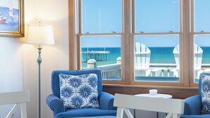 Quarto com uma grande janela com vista para o mar, um quadro emoldurado e duas cadeiras azuis com uma mesa branca.