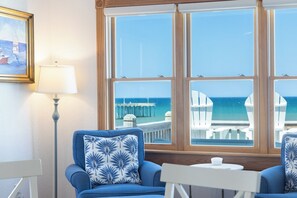 A room with a large window overlooking the sea, a framed painting, and two blue chairs with a white table. - Whit's End: Oceanfront, Private Pool, Elevator, Hot Tub, Theater and Game Room (Nags Head)
