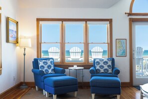 A room with two blue armchairs, a small round table, and a view of the ocean through large windows. - Whit's End: Oceanfront, Private Pool, Elevator, Hot Tub, Theater and Game Room (Nags Head)