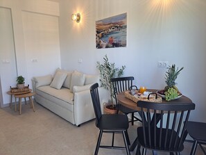 A living room with a sofa, a dining table with chairs, a small side table, and a wall-mounted light fixture. - ALINTHEA 3, Alinda, Leros (Leros)