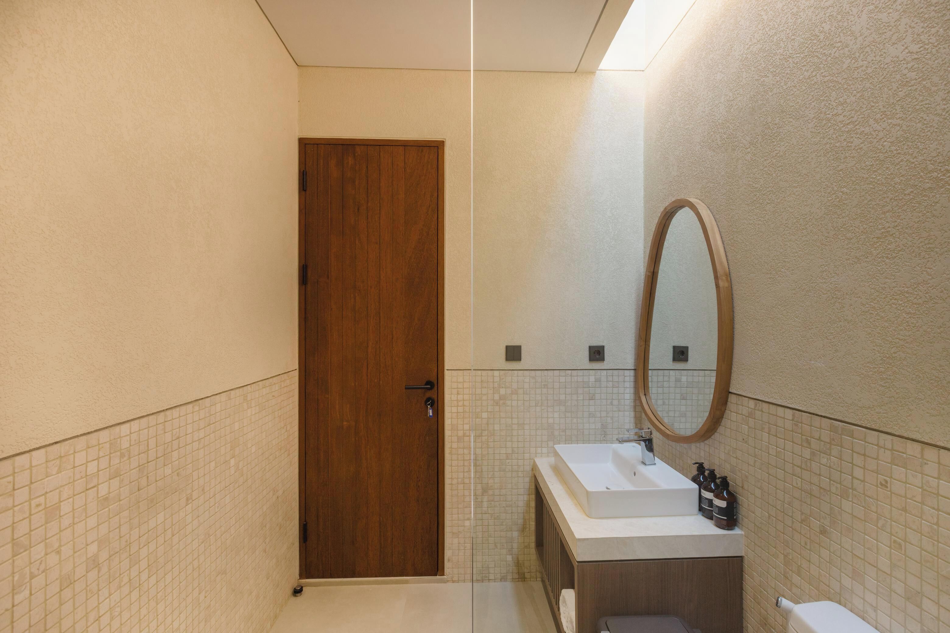A bathroom with a wooden door, a mirror, a sink, and tiled walls.