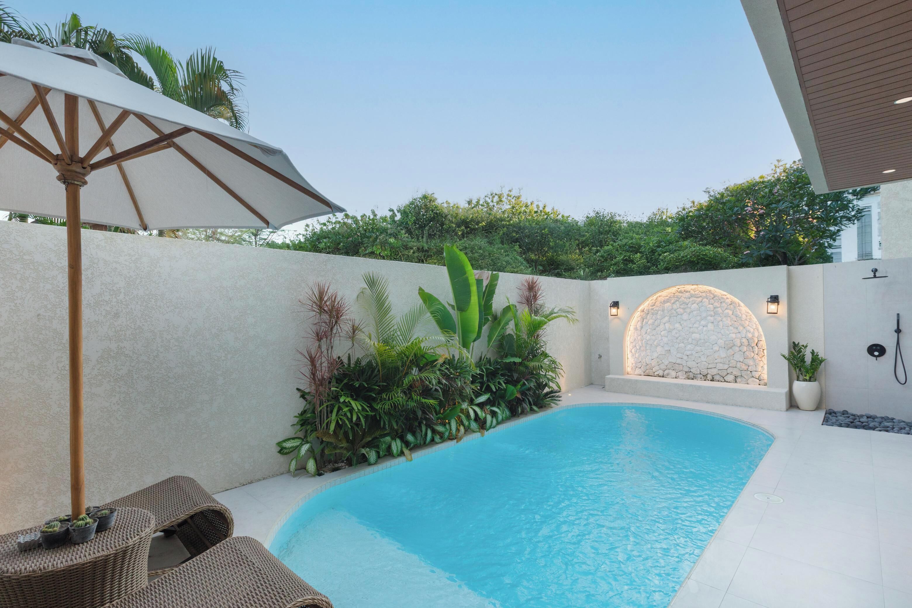 Una piscina con borde de baldosas blancas, rodeada por un muro bajo con plantas y una pared de piedra decorativa. Se observa una silla de mimbre y una sombrilla blanca.