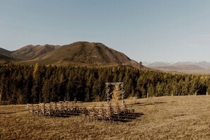 An outdoor wedding setup with a wooden arch and chairs in a grassy field with a forest and mountains in the background.