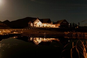 A large, illuminated house with a reflective pond in front, surrounded by hills. - Amazing Log Cabin Rental with Pool Table in the Mountains in West Glacier, Montana (West Glacier)