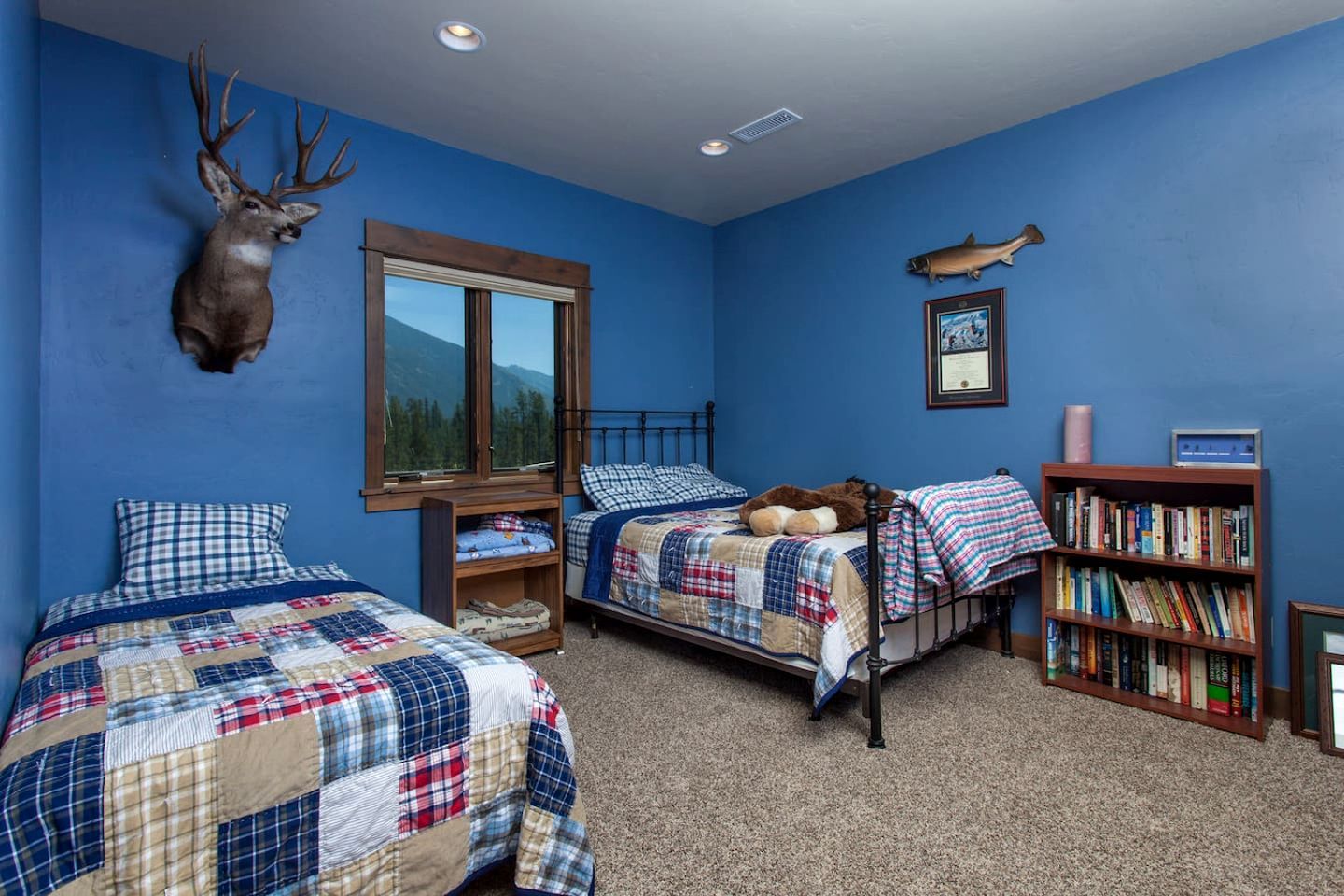 Une chambre avec deux lits, une tête de cerf accrochée au mur, un poisson décoratif, une bibliothèque et une fenêtre donnant sur la montagne.