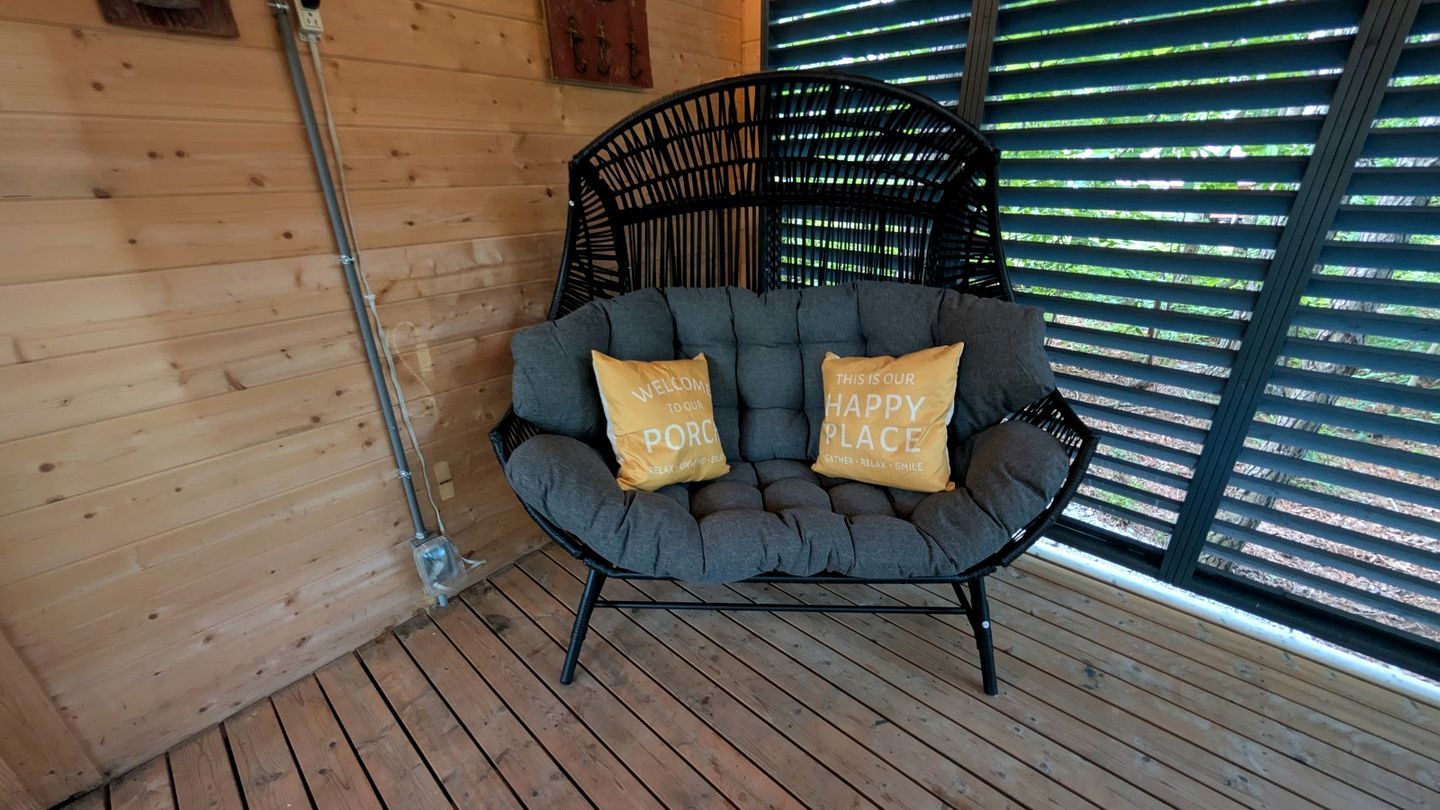 Una silla acolchada gris con dos almohadones, uno con el texto 'WELCOME TO OUR PORCH' y otro con 'THIS IS OUR HAPPY PLACE', ubicada en una terraza de madera con una pared de madera y una barandilla de metal al fondo.