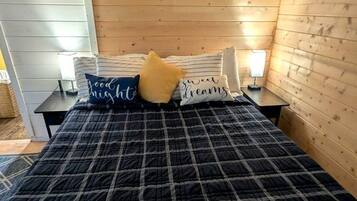 A bedroom with a plaid bedspread, a white headboard, a wall-mounted air conditioner, and a bedside table with a lamp.