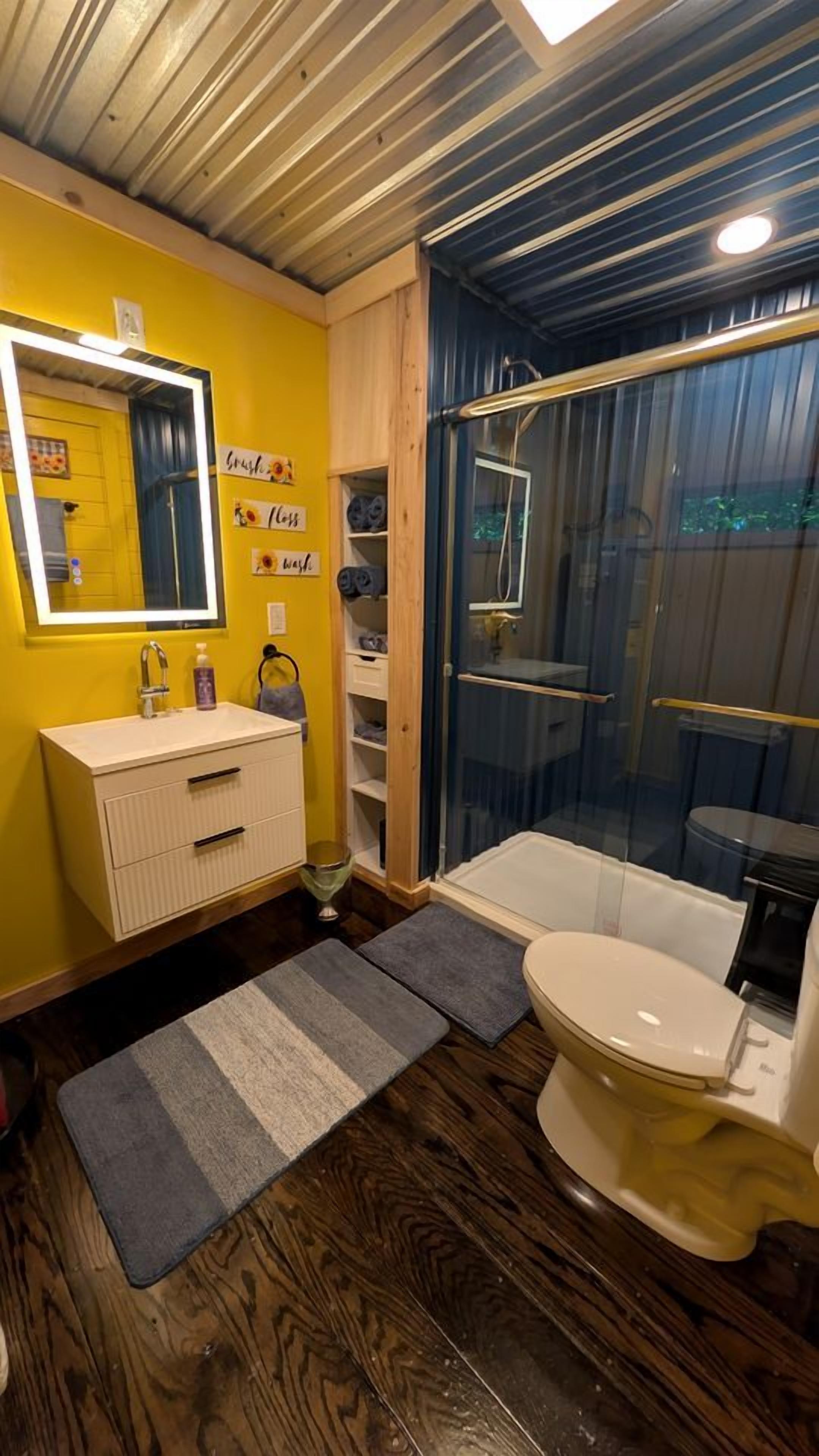 Een badkamer met een gele wand, een witte wastafel, een spiegel, een douchecabine en een toilet.