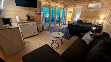 A cozy cabin-style room with a bed, a sofa, a small table, a desk with a coffee machine, and a sliding glass door leading to an outdoor deck.