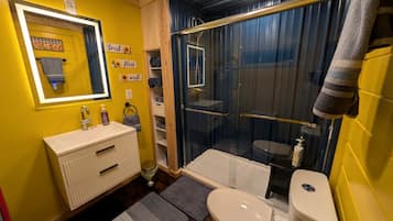 Een badkamer met een gele wand, een witte wastafel, een douche met glazen wanden en een toilet.