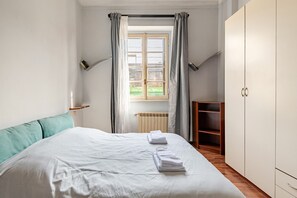 Ein Schlafzimmer mit Bett, Heizkörper, einem Fenster mit Vorhängen, einem Kleiderschrank und einem Regal.