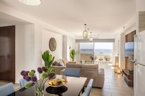 Ein modernes Wohnzimmer mit einer Couch, einem Esstisch und Stühlen, einer Küchenzeile und einem Balkon mit Meerblick.