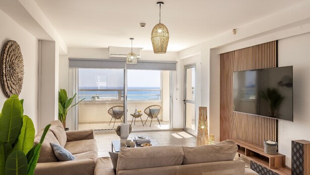 A modern living room with a sofa, TV, and a balcony with a view. - 3 Bedroom Penthouse In the Absolute Center Of Protaras (Paralimni)