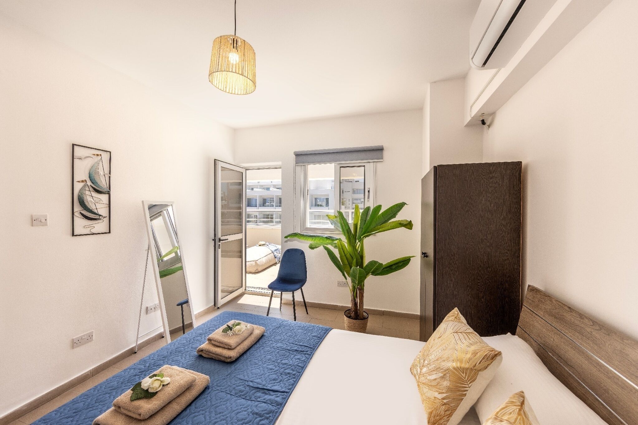Une chambre d’hôtel avec un lit, une grande plante en pot, une chaise, un miroir et un luminaire suspendu.