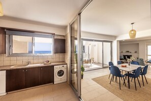 Een moderne keuken met een wasmachine, een eethoek met blauwe stoelen en uitzicht op zee door een glazen deur.