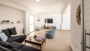 Ein modernes Wohnzimmer mit einer Couch, einem Couchtisch und einem Sitzsack. Ein Fernseher ist an der Wand befestigt und eine Tür führt in einen weiteren Raum.