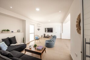 Een moderne woonkamer met een bank, salontafel en een loungestoel. Er is een televisie aan de wand bevestigd en een deur naar een andere kamer.