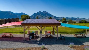 A covered picnic area with tables and chairs, surrounded by a gravel path and a grassy field, with mountains in the background.