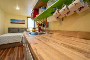 Una pequeña kitchenette con encimera de madera, un lavabo y una estantería con tazas blancas.