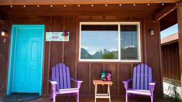 A cabin with a turquoise door, two purple Adirondack chairs, a window with a mountain view, and a welcome sign.