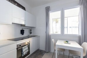 A compact kitchen with white cabinets, a built-in oven, and a window with curtains. - Elianto (Bologna)