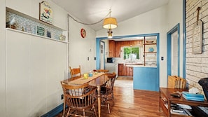 Un comedor con una mesa y sillas de madera, una lámpara colgante, y una cocina visible a través de un vano abierto.