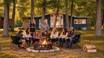 A group of people sitting around a campfire in a wooded area with a small cabin in the background.