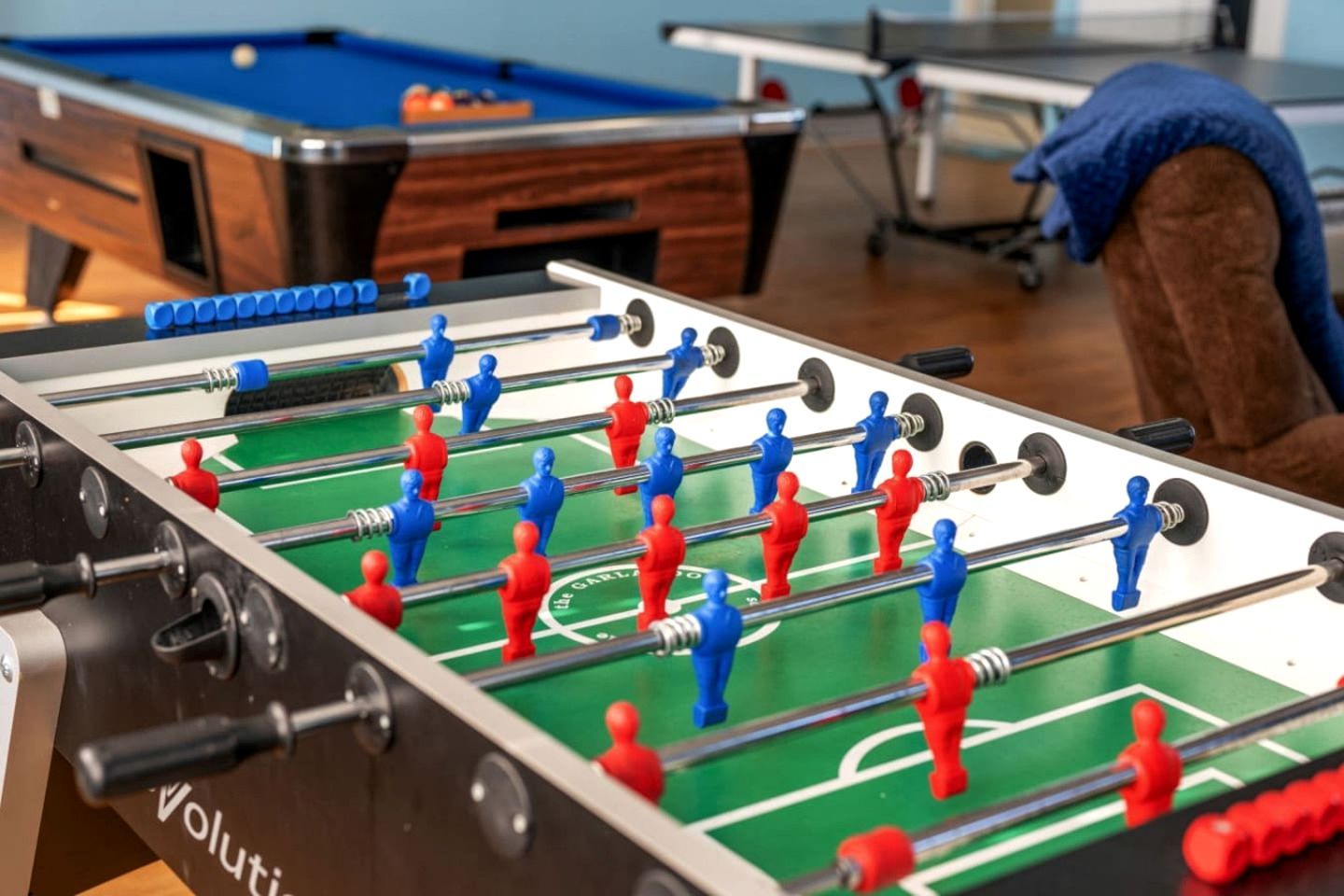 Una taula de futbolí amb jugadors vermells i blaus, una taula de bitlles al fons, i un sofà marró.