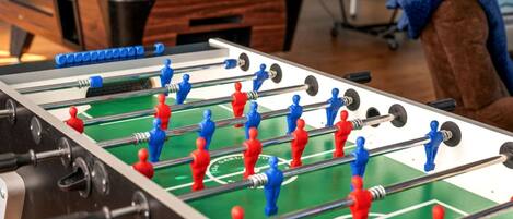A foosball table with red and blue players, a pool table in the background, and a brown couch.