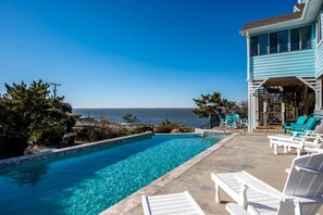 A swimming pool with clear water, surrounded by lounge chairs and a building with a view of the sea. - Gorgeous Beach House with Incredible Water Views for an Unforgettable Escape in Duck, North Carolina (Duck)