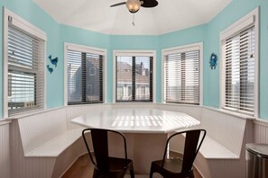 Une pièce avec une table ronde, deux chaises, un ventilateur de plafond et des fenêtres équipées de stores.