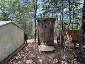 A rustic wooden outhouse with a crescent moon cutout, surrounded by a forest.