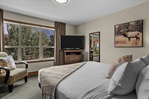 Ein Hotelzimmer mit einem großen Bett, einem Fernseher, einem Schreibtisch und einem Fenster mit Blick auf Bäume und Berge.